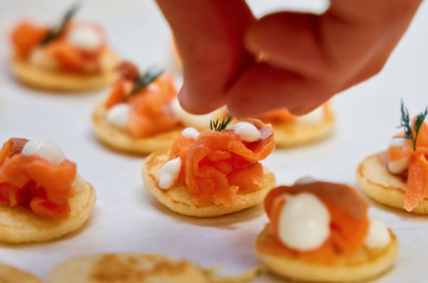 A close-up of a hand delicately garnishing small blinis topped with smoked salmon and a dollop of cream.