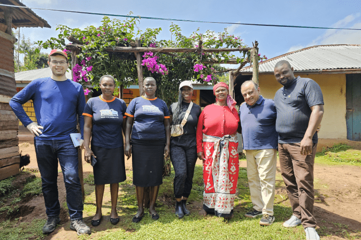 A group of people, including two SAIT students, stand together in a lush outdoor setting in Kenya, with trees and flowers in the background.